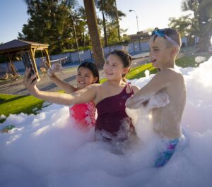 Three children in swimsuits stand in a foam-filled outdoor pool, posing for a selfie with a smartphone.