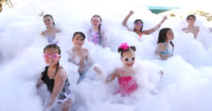 Group of smiling children in swimsuits playing in thick foam at a foam party.